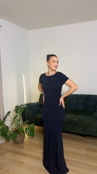 Moteris pozuoja vilkėdama aptemptą tamsiai mėlyną ilgą iki grindų suknelę svetainėje su žalia sofa ir vazoniniu augalu.