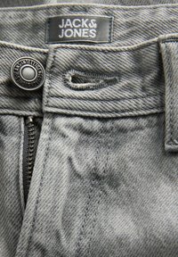 Gros plan sur la ceinture d’un jean en denim gris montrant l’étiquette Jack & Jones, un bouton en métal avec le texte de la marque, une fermeture éclair et les détails de la couture de la boutonnière.