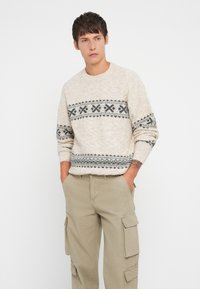 Šviesiai smėlinis megztinis, pagamintas iš tekstūruoto mezginio, su tamsiai pilku geometriniu raštu per krūtinę ir rankoves. Derinamas su chaki spalvos kargo kelnėmis.