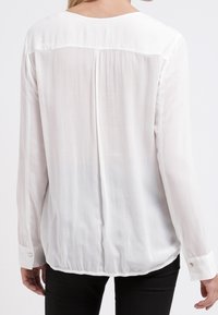 Blouse blanche à manches longues en tissu lisse et léger. Encolure arrondie, poignets boutonnés et détail discret de couture au dos.