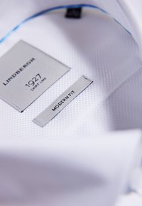 Witte shirt interieur met labels: "Lindbergh," "1927 Shirt Line," en "Modern Fit." Textuurstof met een gladde afwerking. Blauwe accent aan de binnenkant van de kraag.