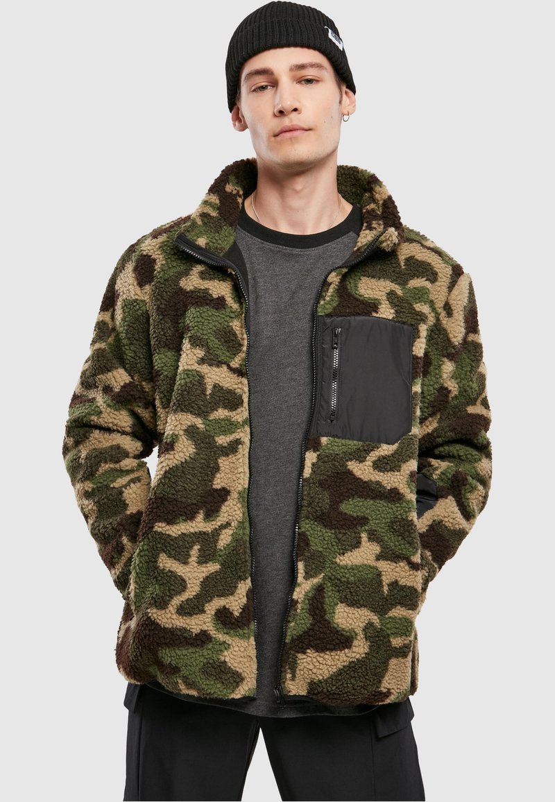 Fleecepaita, jossa on maastokuvio vihreän ja ruskean sävyissä. Siinä on vetoketjullinen rintatasku ja korkea kaulus.