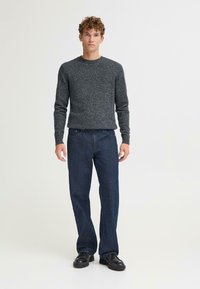 Suéter gris de punto con cuello redondo, combinado con unos vaqueros sueltos de color azul oscuro y zapatos negros, sobre un fondo claro y liso.