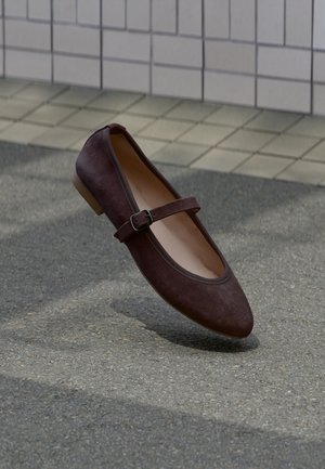 Chaussure plate Mary Jane en daim marron foncé avec bride à boucle, flottant au-dessus d'un trottoir extérieur près d'un mur carrelé.