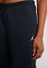 Una mano che riposa vicino a pantaloni sportivi neri con un piccolo logo Jumpman bianco vicino alla tasca e una cintura regolabile con coulisse.
