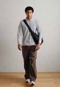 Γκρι μακρυμάνικο πουκάμισο, καφέ παντελόνι, μαύρη τσάντα ώμου, περπατώντας πάνω σε ξύλινο πάτωμα με ουδέτερο φόντο τοίχο. Casual ντύσιμο.