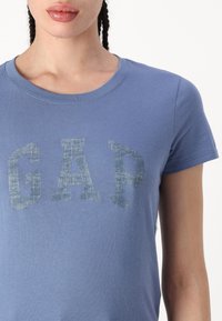 Sinine puuvillane T-särk, millel on kulunud teksatootega "GAP" logo rinnal; ümmargune kaelus ja lühikesed varrukad.