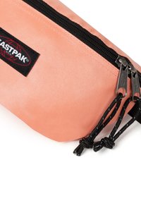 Peachfärgad pennfodral i strukturerad kanvasmaterial, svart dragkedja, dubbla silverfärgade draghandtag och en logotyppatch.