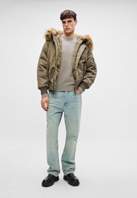 Młody mężczyzna ubrany w oliwkową kurtkę z kapturem i futrzanym wykończeniem, beżowy sweter, jasnoniebieskie dżinsy i czarne buty, stojący na tle jednolitego tła.