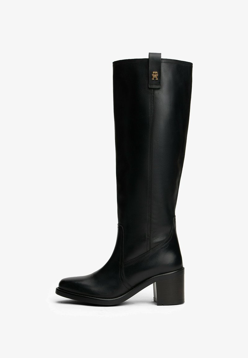 Bota de cuero negra hasta la mitad de la pantorrilla con tacón robusto y emblema dorado en la pestaña de tirador, acabado suave, punta redondeada, costuras laterales y suela ligeramente elevada.