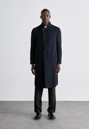 Navy quilted overcoat with a high collar, button front, and side pockets, paired with black trousers and shoes on a neutral background.