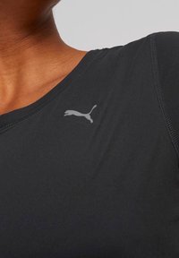 Svart idrottströja med rund halsringning, slät textur och en liten grå Puma-logotyp på bröstet. Närbild av tyget.