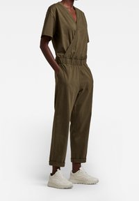 Olivgrön jumpsuit med omslagsdesign, korta ärmar, elastisk midja och avslappnad benpassform. Matchad med vita sneakers.