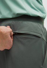 Hand som drar upp en liten dragkedjeficka på mörkgrå byxor som bärs under en ljusgrön skjorta.