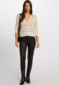 Cardigan lavorato a maglia di crema chiara con motivo a diamante, bottoni neri e maniche lunghe, abbinato a pantaloni neri aderenti e stivali neri lucidi.