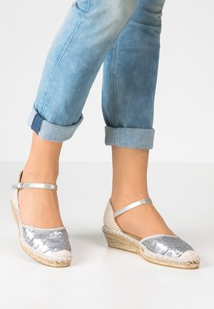 Silver sequined espadrille wedge with a jute sole, rounded toe, and adjustable ankle strap. Denim cuffed pants complement the footwear.
