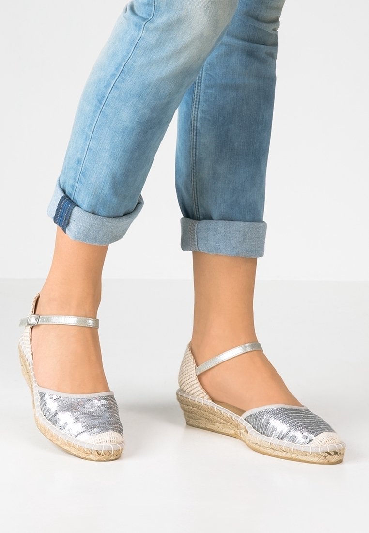 Espadrille compensée argentée à sequins avec semelle en jute, bout arrondi et bride de cheville ajustable. Pantalon en jean retroussé complétant la chaussure.