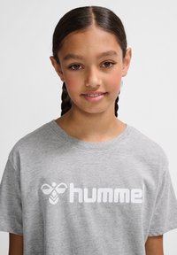 Młoda osoba z splecionymi włosami, ubrana w szary t-shirt z białym logo "hummel", lekko się uśmiecha na jasnym, jednolitym tle.