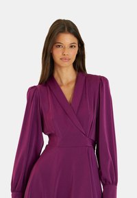 Wickelkleid in tiefem Lila, mit langen Puffärmeln, einem V-Ausschnitt, einer taillierten Passform und einem fließenden Rock. Glattes Material mit dezentem Glanz.