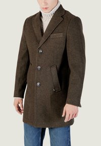 Cappotto in misto lana marrone con rever a punte, quattro bottoni, tasche laterali e una vestibilità sagomata, indossato sopra un dolcevita chiaro.