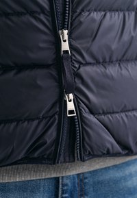 Gros plan d'une doudoune bleu marine avec double fermeture éclair argentée et languettes en tissu, portée sur un pull gris et un jean bleu.