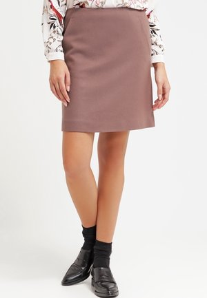 Kvinde iført et malvafarvet knælange nederdel, blomstret bluse, sorte sokker og sorte loafers, stående med krydsede ben på en hvid baggrund.