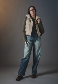 Beige puffer vest over a mørkegrønn tettsittende topp, kombinert med lyseblå vide jeans. Har mørkebrune sko og mønstrede hansker.