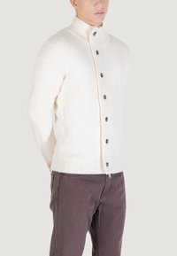 Joven vestido con un cárdigan de punto blanco abotonado y pantalones marrón oscuro, de pie con las manos detrás de la espalda contra un fondo neutral.