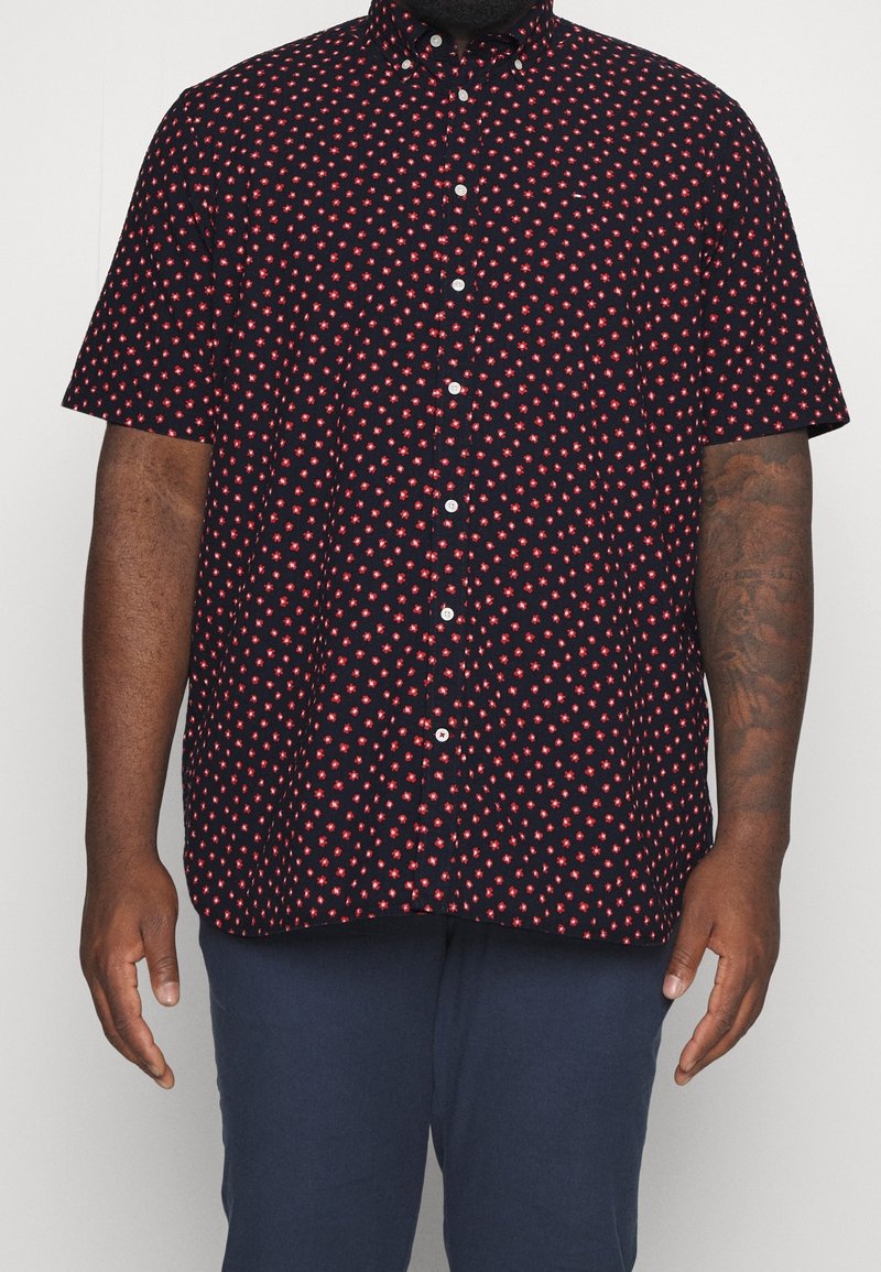 Homme portant une chemise noire à manches courtes avec un motif floral rouge et un pantalon bleu marine, debout devant un fond uni.