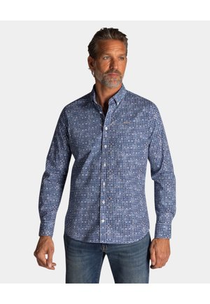 Homme à la barbe grise portant une chemise à manches longues bleu à motifs boutonnée et un jean bleu, regardant légèrement sur le côté.