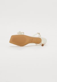 Beige högklackad sandalett med en slät textur, som har en smal klack, justerbara remmar och en dekorativ rosettdetalj på sidan.