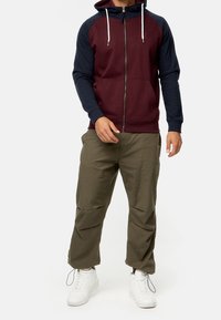 Geverde hoodie in bordeaux en marineblauw, met een capuchon met trekkoord en ribbelmanchetten. Gepaard met olijfgroene cargobroek en witte sneakers.