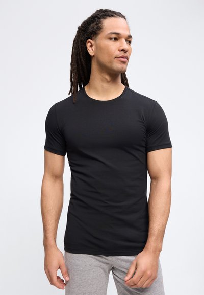 Άνδρας με μακριά dreadlocks που φοράει εφαρμοστό μαύρο κοντομάνικο πουκάμισο και γκρι παντελόνι, κοιτάζοντας προς τα δεξιά σε απλό φόντο.