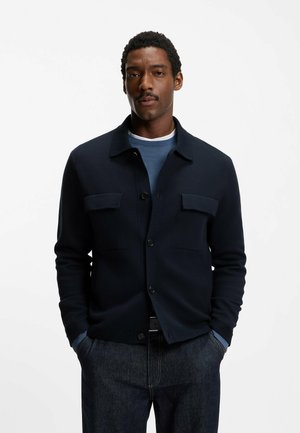 Mand med kort krøllet hår og overskæg iført en marineblå jakke over en blå sweater og mørke jeans, stående med hænderne i lommerne.