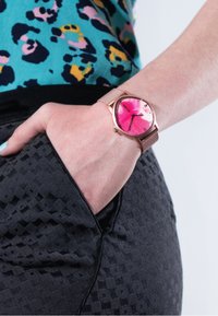 Rund, pink ur med et mønstret urskive, kasse i rosaguld og mesh-rem. Båret på en hånd, der hviler på en sort struktureret talje.