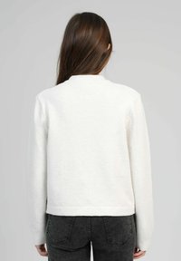 Femme aux longs cheveux bruns portant un pull blanc et un jean noir, vue de dos sur un fond gris uni.