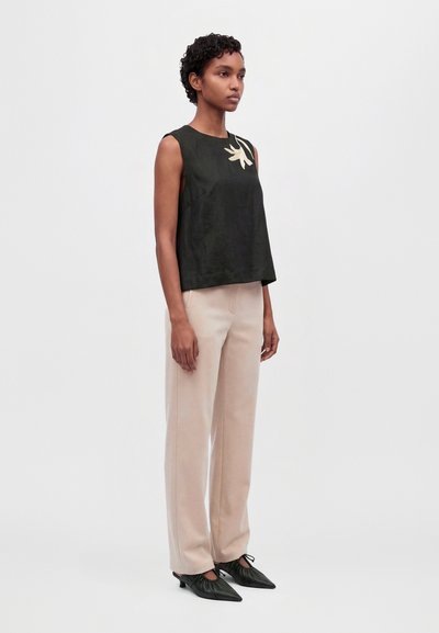 Le modèle porte un haut foncé sans manches avec un détail de fleur crème, un pantalon clair à jambes larges et des chaussures plates noires à bout pointu, devant un fond de studio uni.