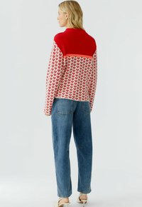Strickpullover mit einem roten oberen Teil, einem weißen Körper und einem sich wiederholenden roten Muster, kombiniert mit hellblauen geraden Jeans.