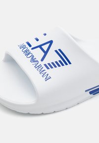 Vit halksand i slät gummi med en blå logotyp med ränder och texten "EMPORIO ARMANI." Öppen tå med rundad form.