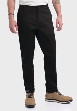 Mann trägt schwarze gerade geschnittene Hose mit beigen Schnürstiefeln, steht mit locker hängenden Armen an den Seiten vor einfarbigem Hintergrund.