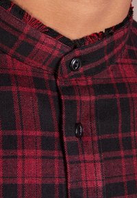 Rode en zwarte geruite blouse met een mandarinelapel, zwarte knopen en licht-uitgefranste randen bij de halslijn en de knopenlijst.