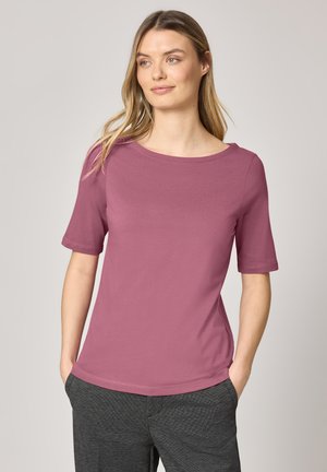 Junge Frau mit hellbraunen Haaren, die ein kurzärmliges mauvefarbenes Shirt und dunkelgraue Hose trägt, die Hände in den Taschen, schaut seitlich weg.