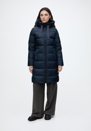 Femme portant un manteau matelassé noir mi-longueur aux genoux avec un col montant et des cordons de serrage, pantalon large foncé et chaussures noires, debout face à l’avant.