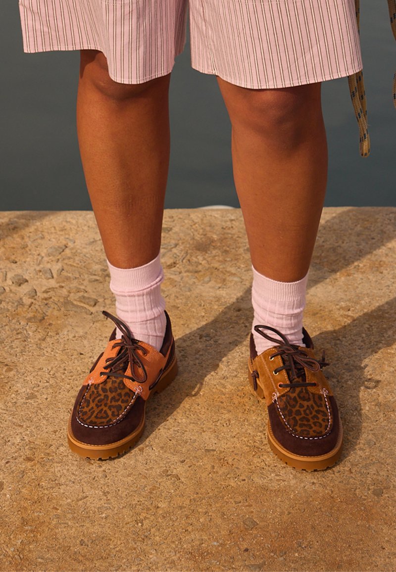 Jambes portant un short rose à rayures, des chaussettes blanches et des chaussures marron à lacets avec des panneaux à motif léopard, debout sur un sol texturé.