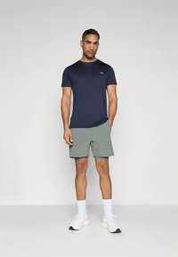 Tmavě modrá košile s krátkým rukávem v kombinaci s světle zelenými šortkami. Model má na sobě bílé sportovní boty a ponožky s jednoduchým designem.