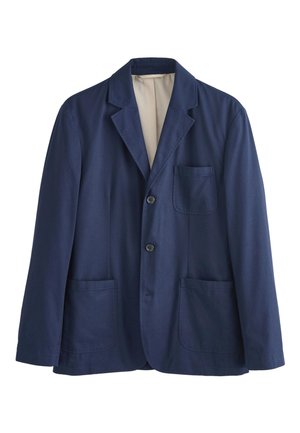 Ung mand med krøllet hår iført en navy blazer og matchende bukser over en hvid t-shirt, hænderne i lommerne, kigger til siden.
