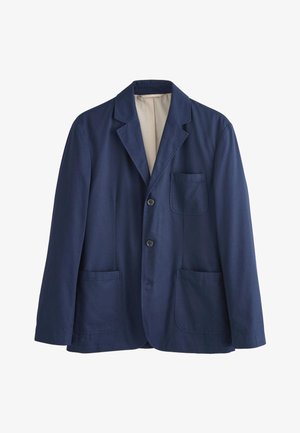 Ung mand med krøllet hår iført en navy blazer og matchende bukser over en hvid t-shirt, hænderne i lommerne, kigger til siden.