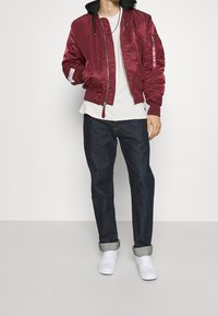 Giacca bomber bordeaux con chiusura a zip, cappuccio nero, polsini a coste e tasche laterali. Abbinata a jeans scuri in denim e sneakers bianche.
