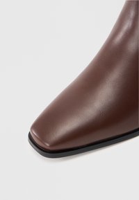 Stivale alla caviglia in pelle marrone con punta affilata, texture liscia e una sottopiede nero sottile. Presenta accenti minimi e linee pulite.
