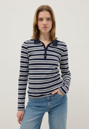 Jonge vrouw staat met hand in de zak, draagt een marineblauw-wit gestreept poloshirt met lange mouwen en lichtblauwe jeans tegen een effen achtergrond.